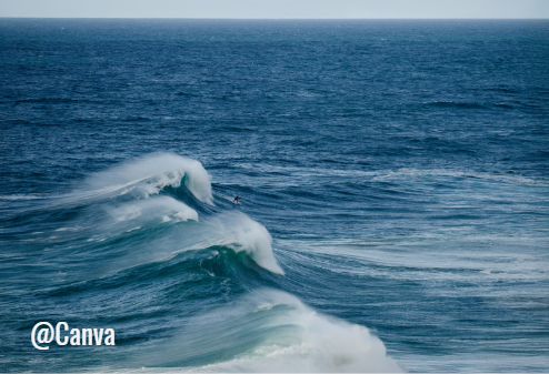 Vue large de l'océan avec de grosses vagues déferlantes formant des crêtes blanches sur une mer d'un bleu profond. On aperçoit une petite silhouette sur l'une des vagues ; watermark "@Canva" en bas à gauche.