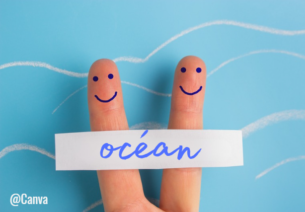 Deux doigts levés décorés de visages souriants dessinés en bleu, tenant une bande de papier sur laquelle est écrit "océan" en écriture cursive. Fond bleu clair avec des traits blancs ondulés suggérant des vagues ;
