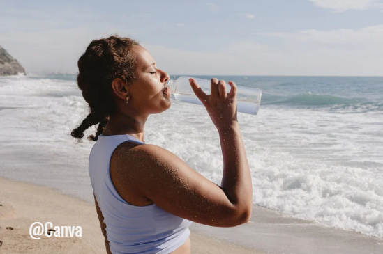 Une femme se tenant sur une plage regarde l'horizon en buvant dans un gobelet transparent, cheveux attachés et vêtue d'un débardeur clair